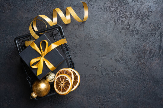 Black gift box with gold ribbon and Christmas ornaments in a basket on dark grunge background