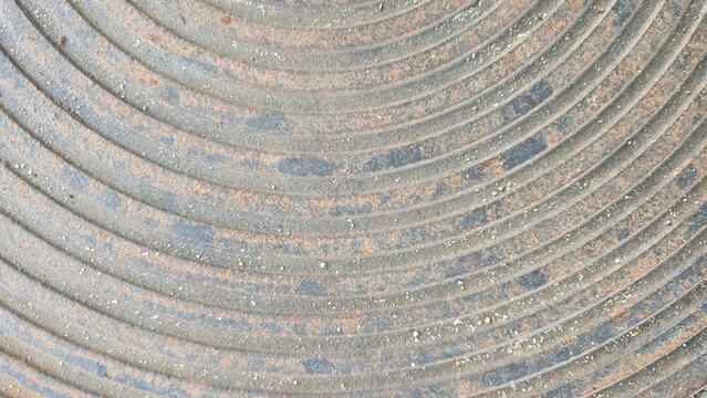 Old cast iron manhole cover with concentric circular pattern. Grunge industrial metal background with curved lines. Close-up of weathered metallic manhole texture showing circular ridges and rust.