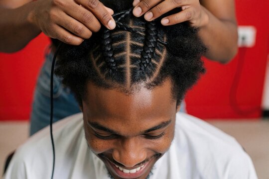 Top view of smiling young afro man getting hair braided by professional stylist. Black male client enjoys hair care service in salon. Skilled hands create neat cornrow braids on head for trendy