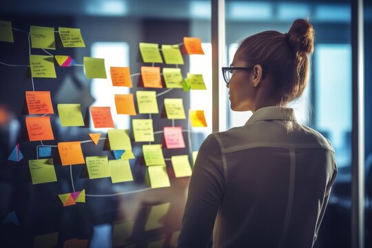 Woman standing in an office, focusing on a transparent board covered with various colorful sticky notes connected by lines, indicating a complex project management or brainstorming session