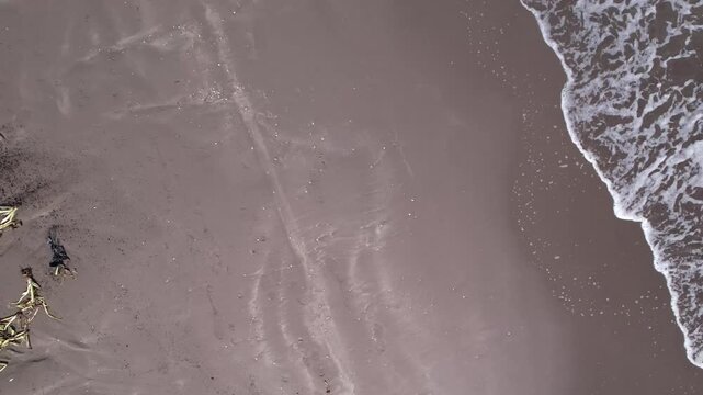 Top down view of brown wet beach sand meeting a foamy sea edge, revealing thin winding water marks across the tidal shore, where waves reach the sand.