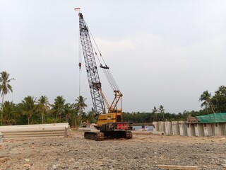 Heavy crawler crane operating at an outdoor infrastructure construction site near a riverbank, surrounded by concrete beams, palm trees, and an Indonesian flag flying on the boom peak. © MUMTAZUN