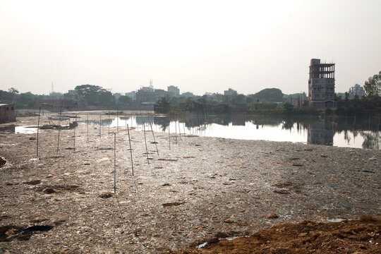Hazy view of a dried polluted tannery lagoon with bamboo stakes and a tall unfinished concrete building beyond, Hazaribagh, Bangladesh