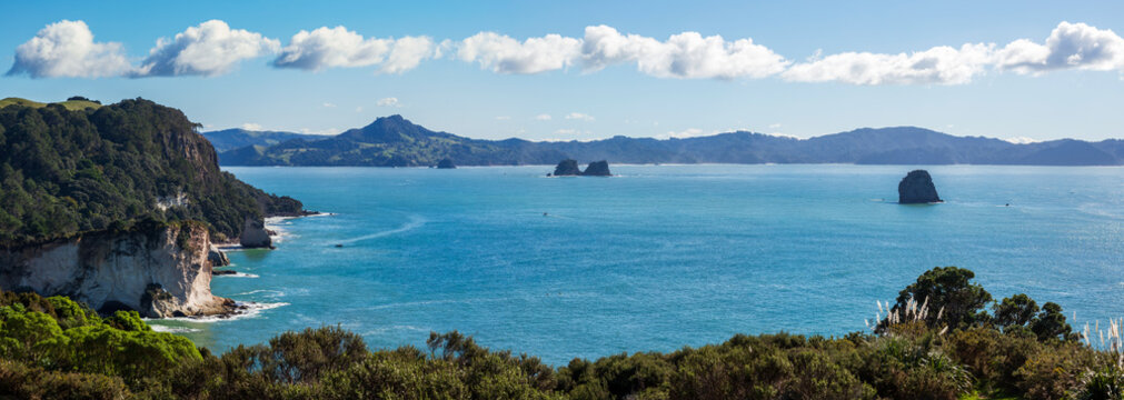 Rugged white cliffs covered in dense greenery rise above the deep blue ocean in Coromandel, with rocky sea stacks and distant hills completing the scenic coastal view