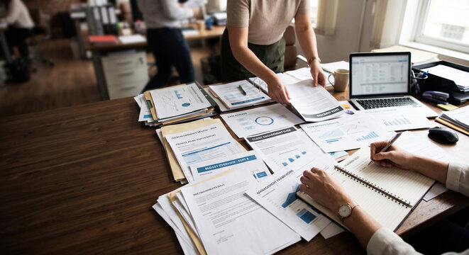 A team of office workers reviewing documents, business charts, and data reports on a busy wooden desk with a laptop