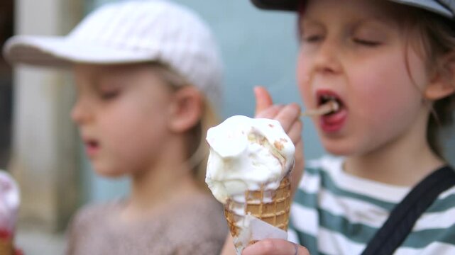 Young girl enjoys spoonful of ice cream beside another child holding cone. Her contented expression suits nostalgic childhood treat, as children savor simple outdoor snack together.