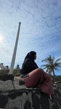 Low-angle silhouette portrait of a woman in hijab sitting on a stone wall by a palm tree, looking out under a dramatic, textured cloudy sky with the sun visible.