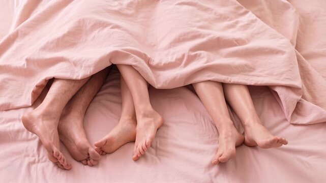 Intimate scene showing bare feet of a couple under soft pink bedding, suggesting comfort and closeness in the bedroom. Three people in a bed