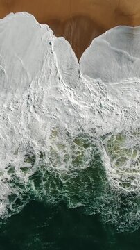 Stunning aerial perspective capturing the immense power of nature as enormous turquoise waves with churning white foam relentlessly crash upon the golden shores of nazare, portugal