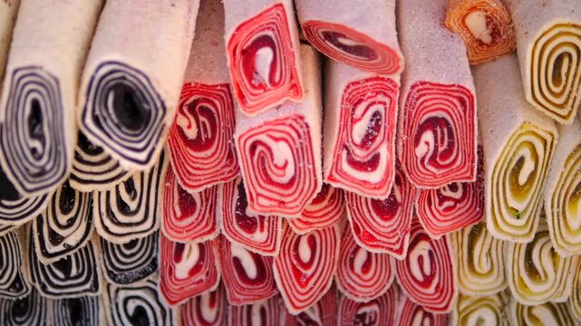 Rows of striped wafer biscuits create vivid closeup, their rolled layers showing red, dark and cream swirls. Colorful cookies hang together in dense display with occasional golden pieces.
