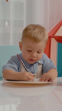 Vertical Screen: A cheerful boy enjoys drawing with colored pencils at a bright table. Colorful toys surround him while he focuses on his creative artwork.