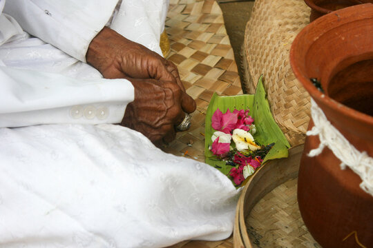 Close-up of an elderly priest hands in white sleeves beside flower offerings on a banana leaf and a clay pot, Ubud, Indonesia