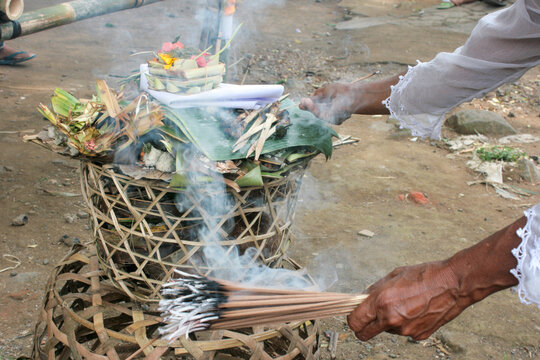 A hand in a lace sleeve waves smoking incense sticks over a woven basket of offerings during a cremation ceremony, Ubud, Indonesia