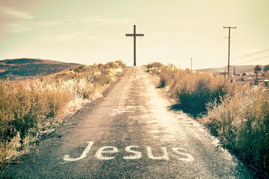 A paved road ascends towards a large dark cross with the word Je