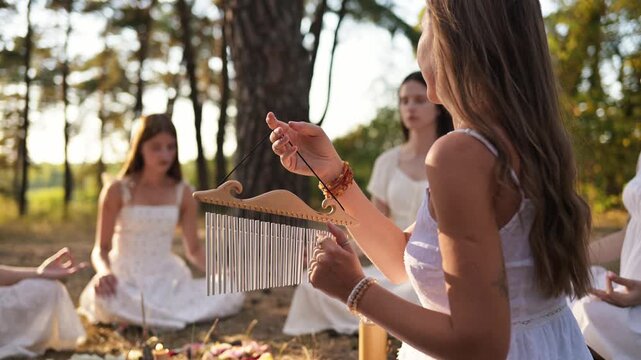 Women in white dresses engage in a nature gathering, one holding a wooden instrument while others sit nearby among flowers and pine trees