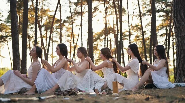 Women in white dresses form a circle in a forest, braiding hair together, with sunlight streaming through trees and a natural ground surface visible