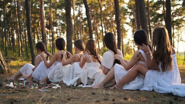 Group of women in white dresses sitting in a circle, braiding each other's hair in a forest setting, surrounded by nature and sunlight filtering through trees