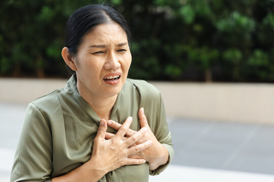 Middle aged 50s Southeast Asian woman experiencing heart pain outdoors, hand on left chest and distressed expression