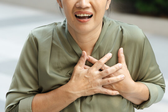 Middle aged 50s Southeast Asian woman experiencing heart pain outdoors, hand on left chest and distressed expression, hand and body part closeup shot