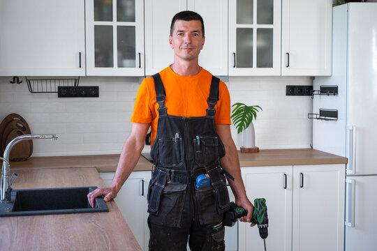 Professional male assembler in orange t-shirt holding a drill machine in kitchen environment, furniture assembly, professional service.