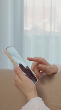 Vertical Screen: A woman sits on a couch at home, holding a smartphone. She swipes on the screen while engaging with social media and connecting with friends.