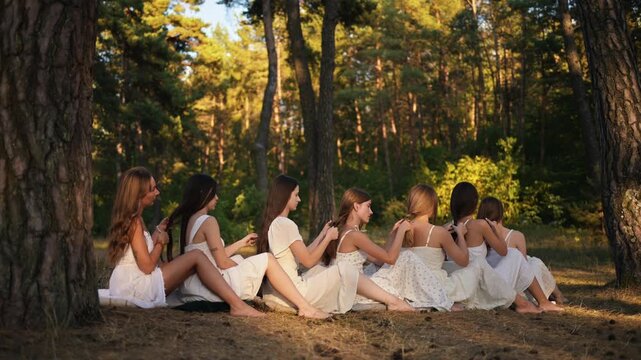 Young women in white dresses sit on the ground in a forest, braiding each other's hair, with tall trees and sunlight filtering through the leaves