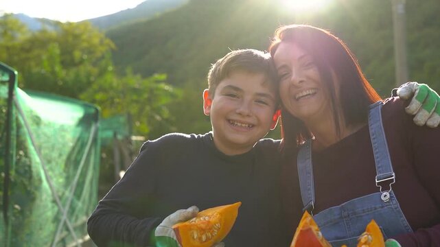 Happy mother and teenager son having fun harvesting organic pumpking during fall season at house vegetable garden while smiling on camera - Farmer lifestyle, family people and autumn season concept