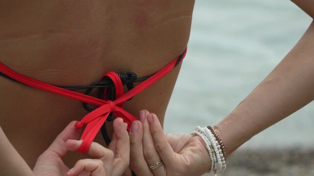 Coastal swimwear adjustment by woman tying red strings on beach shore wearing beaded bracelets. Close-up static shot.