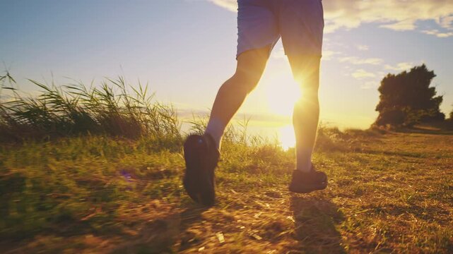 A senior adult is jogging along a grassy path by the water at sunset, wearing blue shorts and black running shoes. The warm glow of the sun highlights their active, healthy lifestyle.