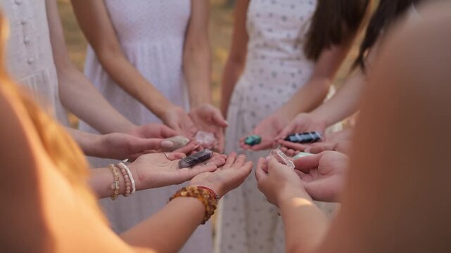 Women in white dresses form a circle, each holding various colorful crystals, engaged in a meditation session outdoors with natural light