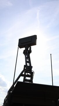 Vertical screen Military radar antenna mounted on an elevated mobile platform stands against bright sunlight with dramatic sun rays and lens flare during outdoor defense display