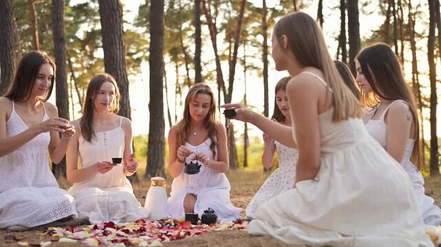 Women in white dresses sit in a circle on the ground, surrounded by flower petals and small cauldrons, engaged in a ritual in a forest during sunset