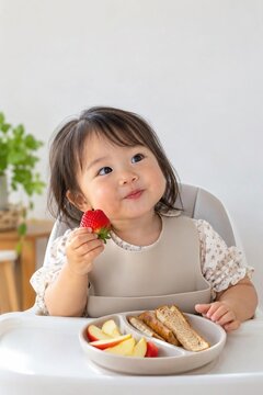 Cute asian toddler girl eating strawberry in high chair with silicone bib with plate of apple with toast, baby led weaning