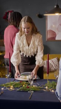 Vertical video of young adult Caucasian woman setting Christmas dinner table with diverse friends at home. Clip suitable for holiday marketing and festive celebration campaigns