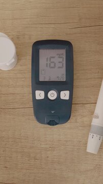 Vertical Screen: A glucometer rests on a wooden table showing a blood sugar level of 163 mg dL. This indicates a very high sugar level related to diabetes management.