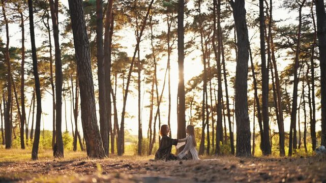 Two women are seated on a blanket in a forest, conversing as the sun sets, casting warm light through the trees surrounding them