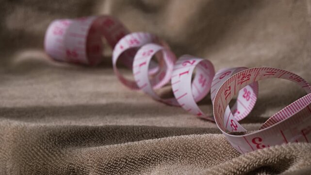 A flexible white measuring tape with red markings lies coiled on a piece of soft textured brown fabric in soft light.