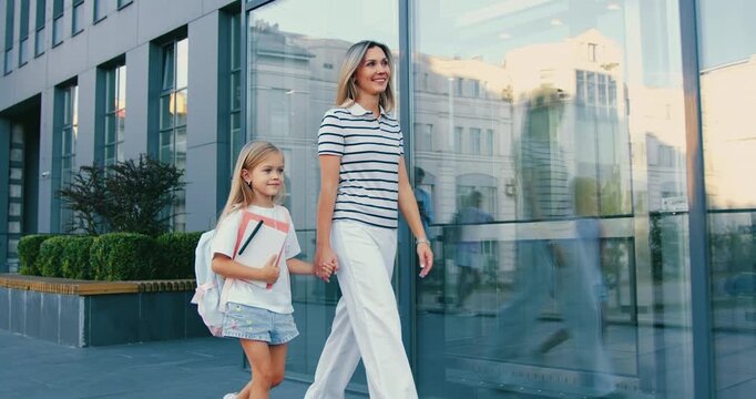 Mother and daughter walking hand in hand past glass building on sunny urban sidewalk, mother smiling and daughter carrying backpack, casual summer clothing and warm daylight reflection scene. Back to