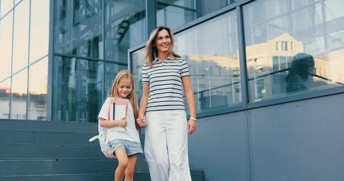 Loving mother leading little daughter to school and entering together with other primary school students. Preadolescent schoolgirl with backpack going to school holding mother's hand. Back to school.