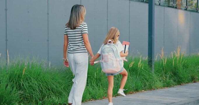 Back view. Mom and daughter walking along path through green park, holding hands in casual summer clothes, lampposts and trees lining walkway, candid rear view capturing gentle bond and relaxed