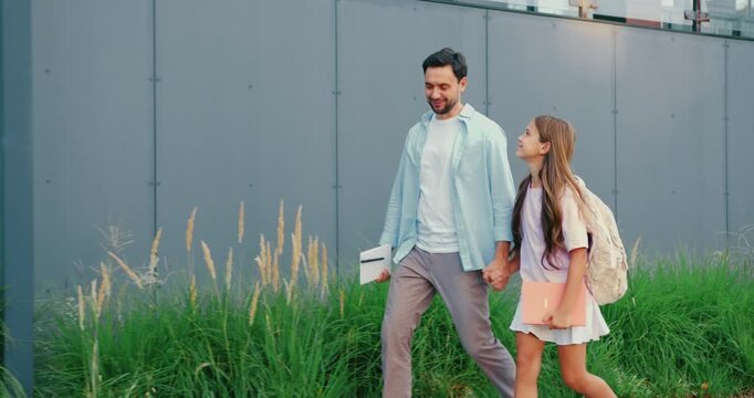 Back view. Father and daughter walking along past glass building on sunny urban sidewalk, holding hands in casual summer clothes, lampposts and trees lining walkway, candid rear view capturing gentle
