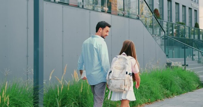 Father walking to school with daughter holding hands on urban sidewalk past concrete wall and greenery, casual daylight family stroll toward school or city building. Back to school.