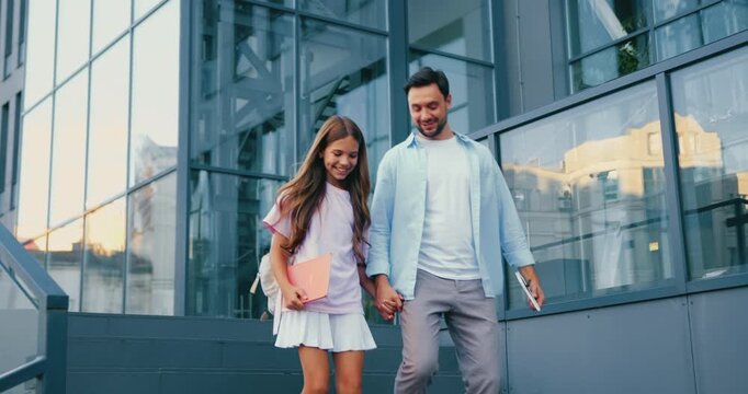 Father and daughter walking hand in hand down urban steps outside modern glass school building, smiling and playful in casual daytime clothes, low viewpoint capturing motion and family connection