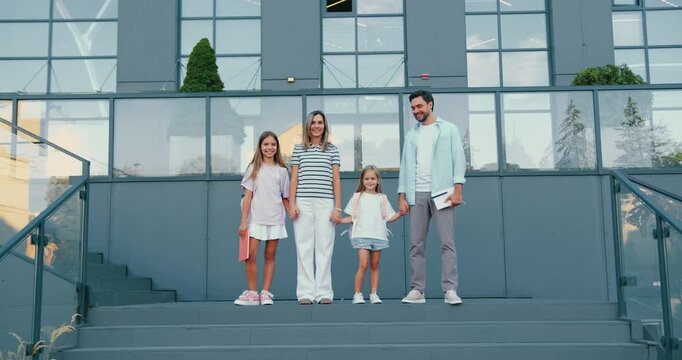 Family of four descending steps outside modern glass building in casual summer clothing looking at camera smiling and holding hands in relaxed urban setting with symmetrical composition and soft