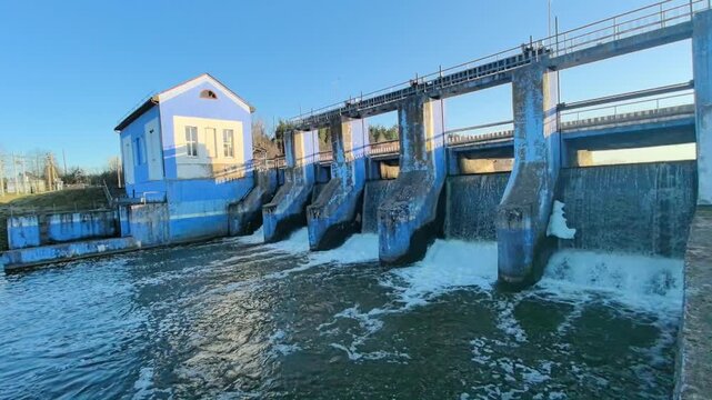 Water is released from a dam into the river below. The structure is located in a rural area and the building next to the dam has a blue and white design. Sunlight shines on the water, creating a brigh
