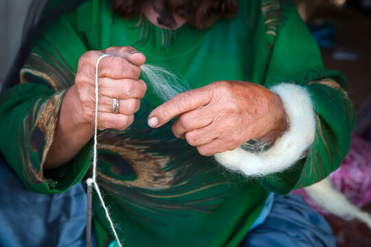Close-up of weathered hands of a Qashqai woman in a green peacock pattern dress spinning white wool with a spindle, Semirom, Iran