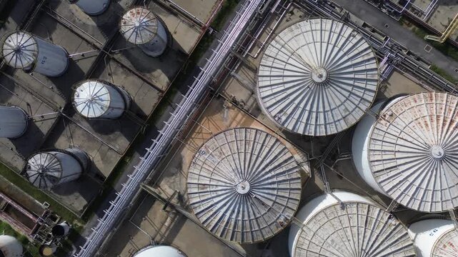 4K drone footage moving sideways high over a port oil depot, looking down. Round tank roofs with radial ribs, smaller tanks in bunds and pipeline rack pass beneath. Original unedited video.