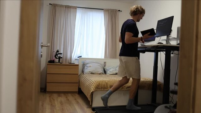 Young man runs on a treadmill while studying programming on a computer and reading a book combining physical exercise with online learning and productivity in his home office