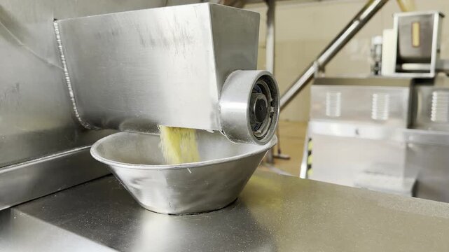 Coarse yellow powder flows from a machine outlet into a metal bowl during industrial ingredient processing