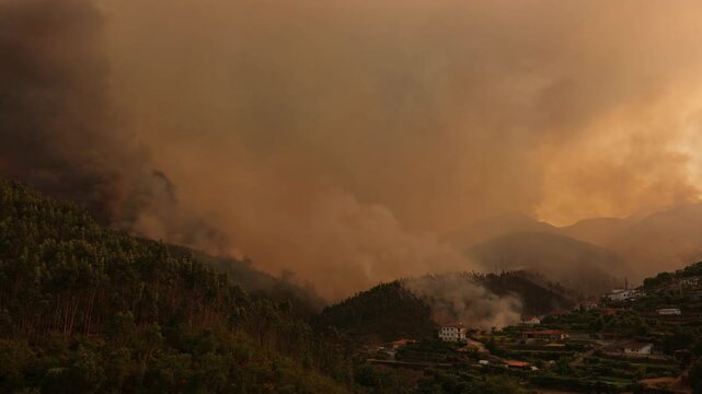 Firefighting helicopter deploying water over a blazing wildfire, dense smoke engulfing forested hills and a vulnerable village during an intense summer heatwave.
